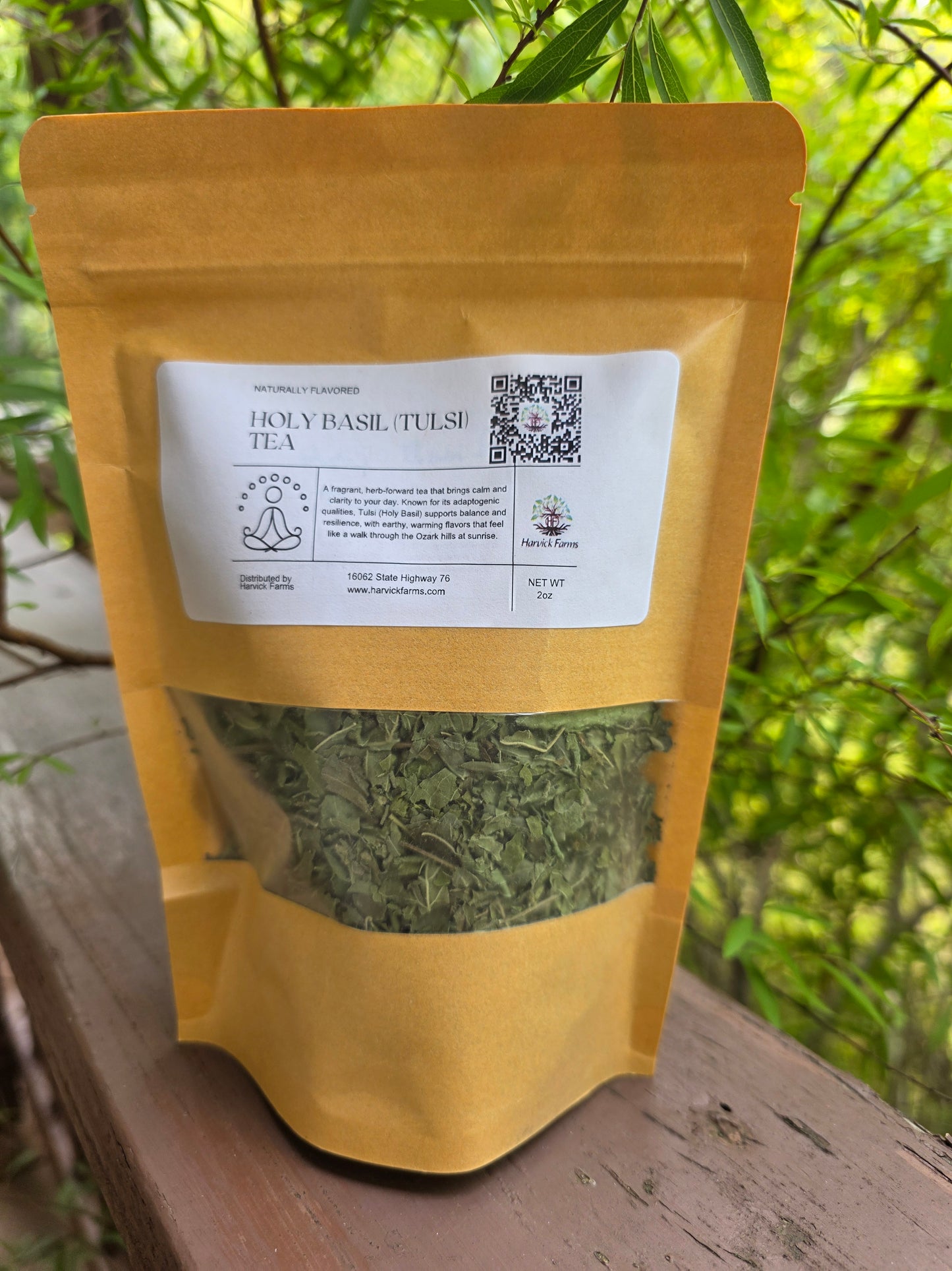 A stand-up pouch of Holy Basil Tulsi Tea placed on a wooden surface outdoors, with green foliage blurred in the background. The pouch is mustard yellow with a white label and a clear window showing dried green tea leaves inside.