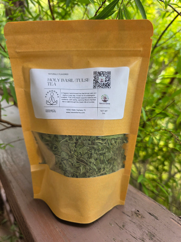A stand-up pouch of Holy Basil Tulsi Tea placed on a wooden surface outdoors, with green foliage blurred in the background. The pouch is mustard yellow with a white label and a clear window showing dried green tea leaves inside.